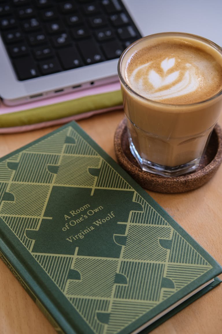 A latte art coffee next to Virginia Woolf's book and a laptop on a wooden desk.