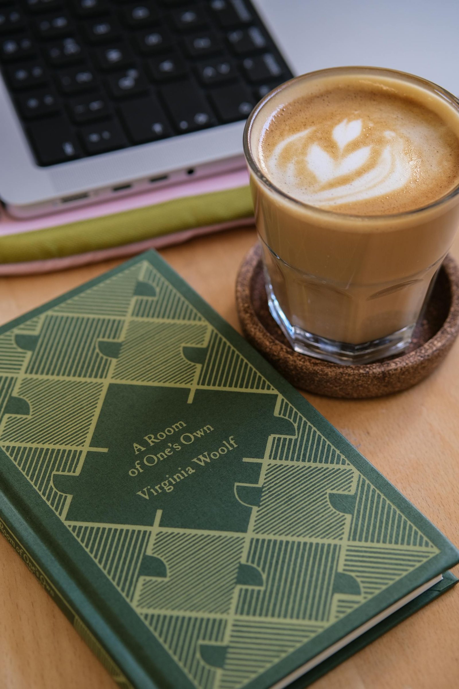 A latte art coffee next to Virginia Woolf's book and a laptop on a wooden desk.