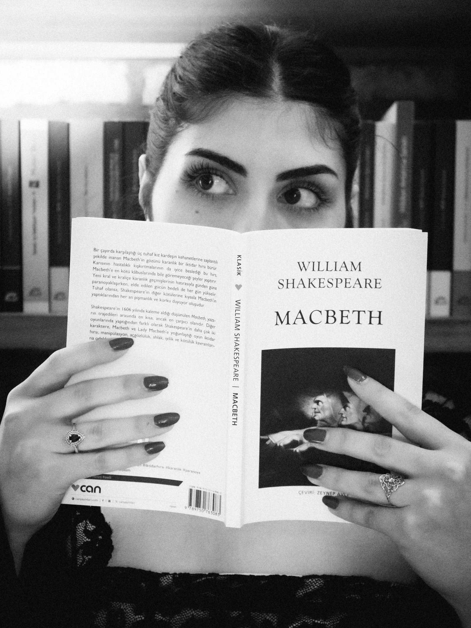 Black and white photo of a woman reading Macbeth by William Shakespeare in a library.
