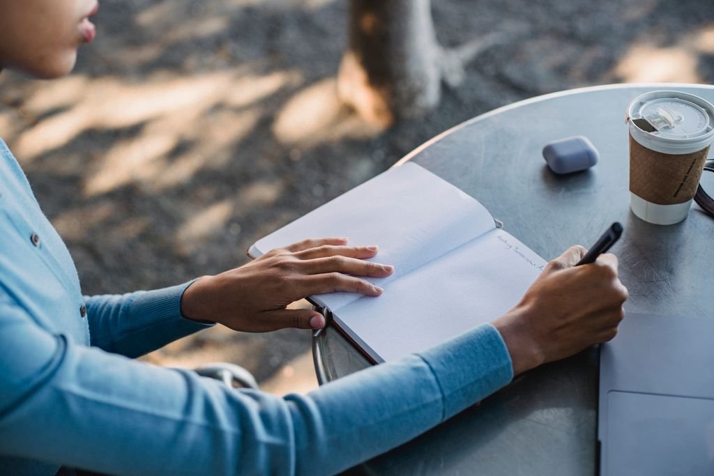 Dialogue Journal - A close-up of a person writing notes in a notebook at an outdoor cafe table with a coffee cup.