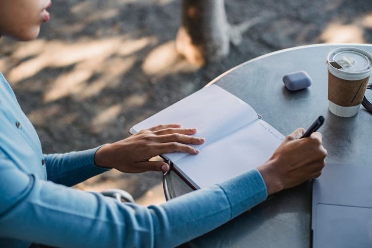A close-up of a person writing notes in a notebook at an outdoor cafe table with a coffee cup.