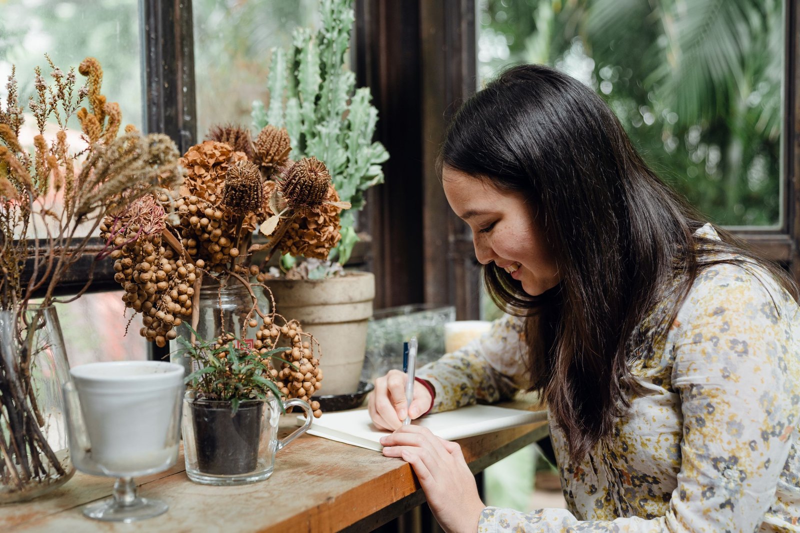 Side view of cheerful young Asian female writer with dark hair smiling and getting inspired while creating in cozy greenhouse