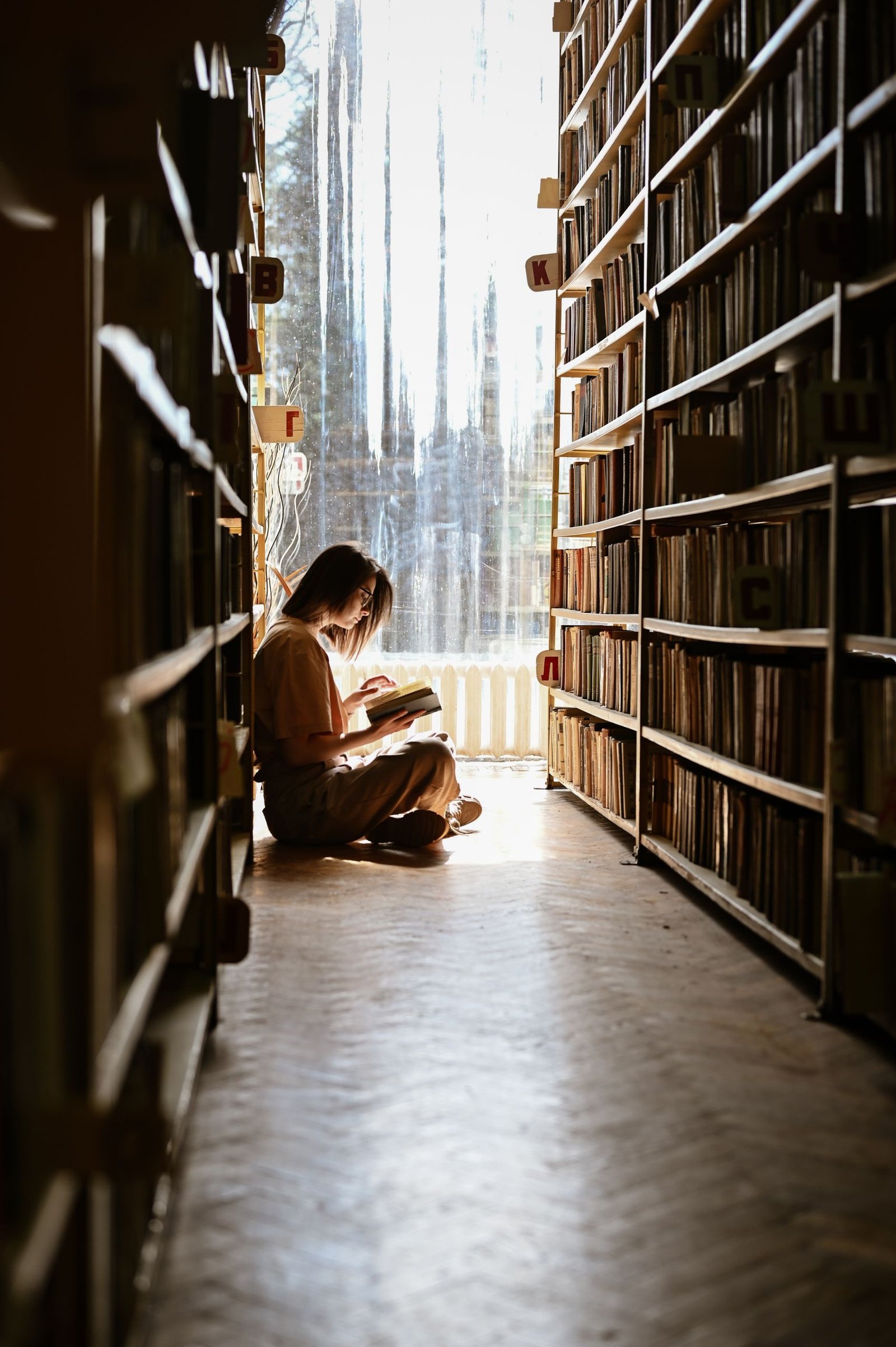 Offline Hobbies - Young girl reading by herself in a serene library with sunlight streaming in.