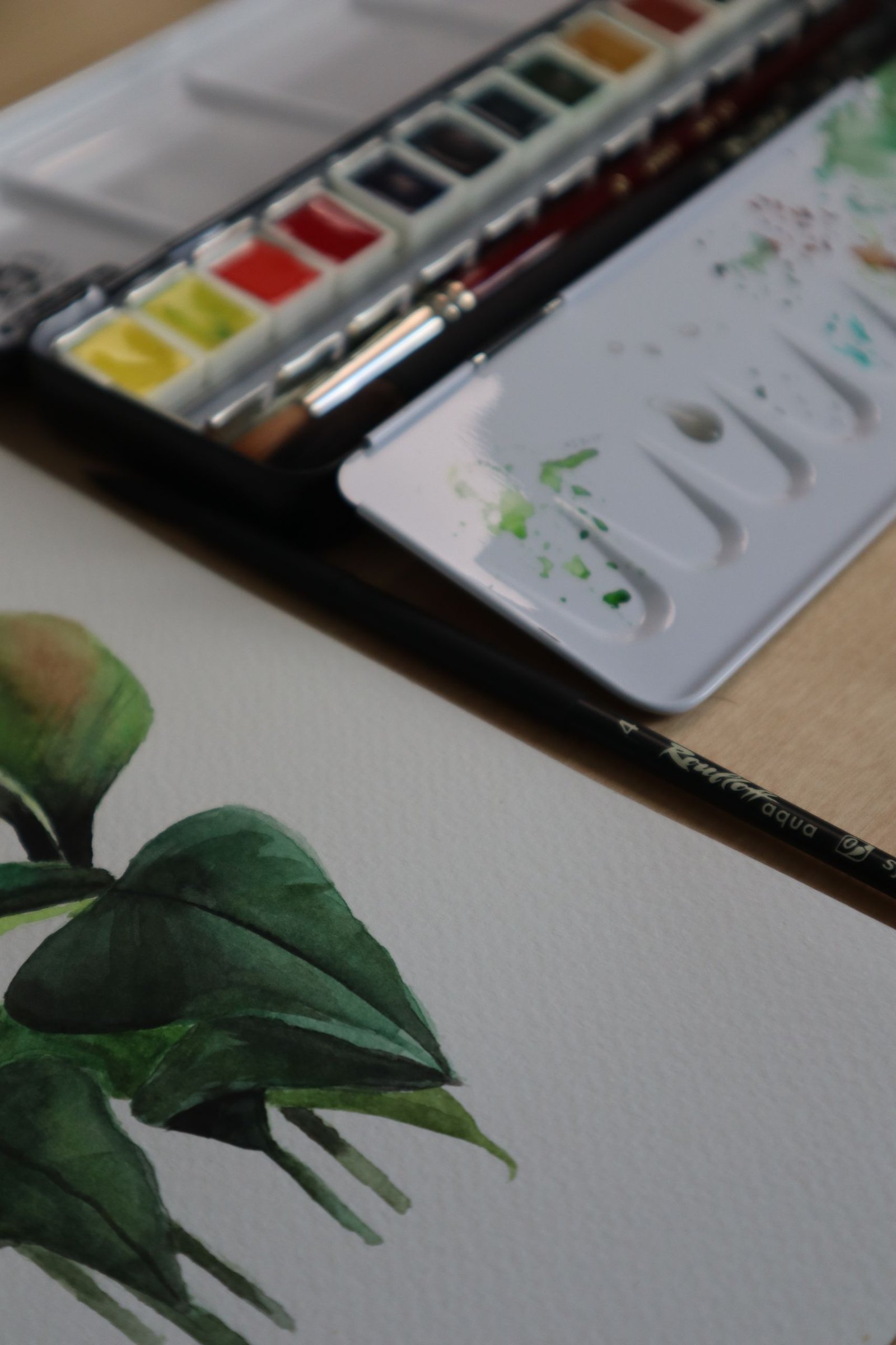 Artistic setup with watercolor paints and brush on wooden surface, focusing on a leaf painting.